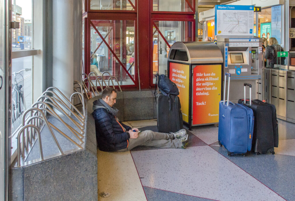 A man sitting on the floor of a train station because the ledge behind him is covered in metal barriers. 